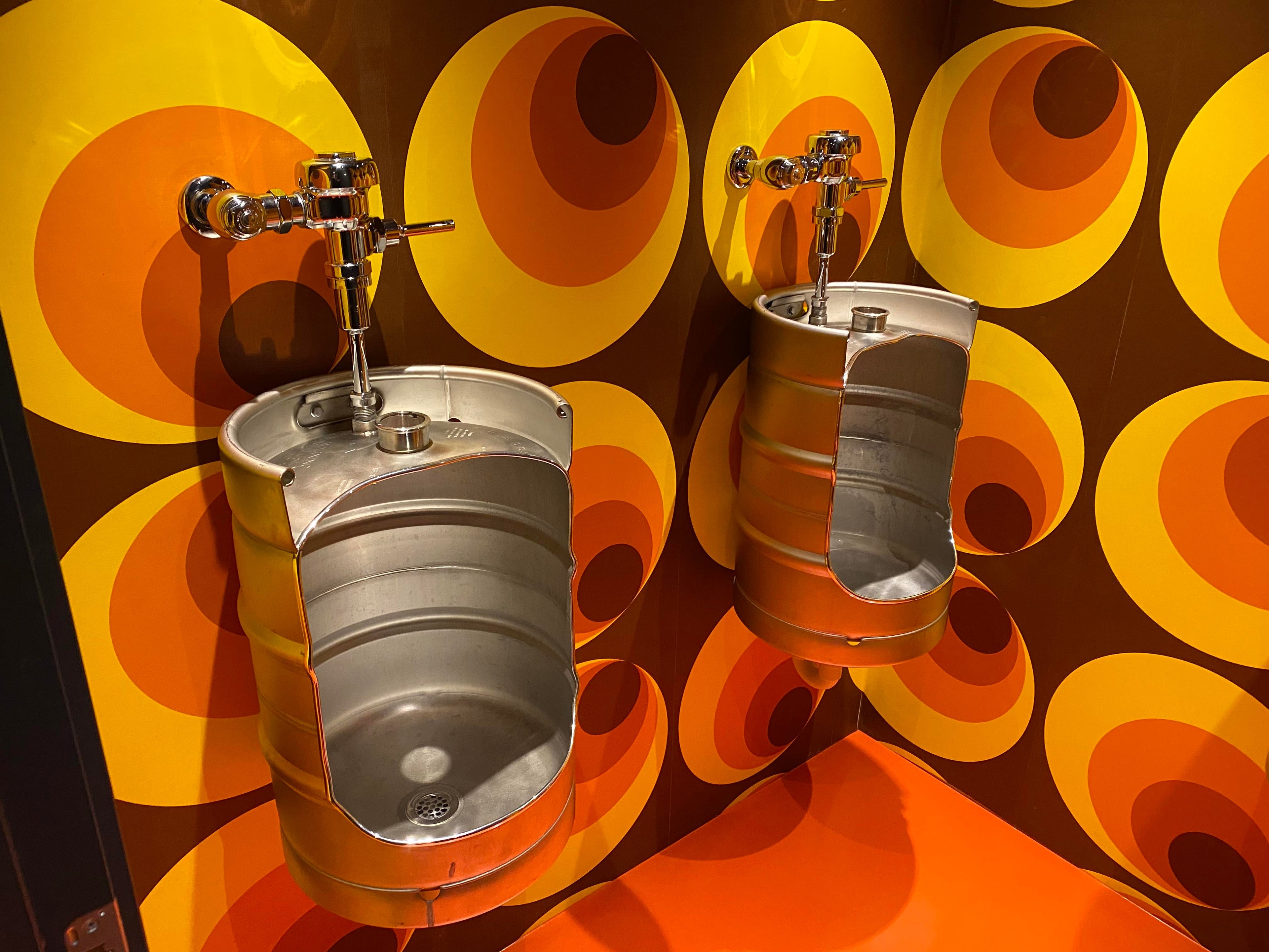 Two metal keg urinals mounted on a wall with orange and yellow circular patterns.