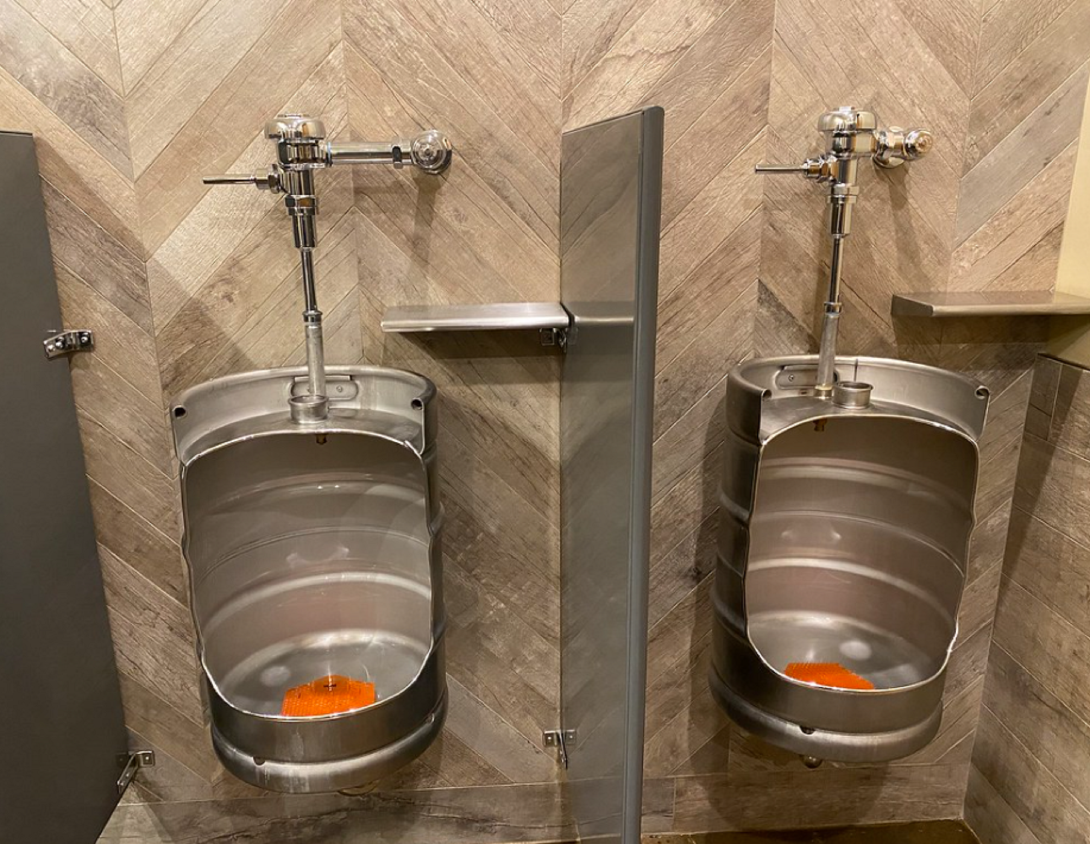 Two keg urinals mounted on a tiled wall with a metallic divider.